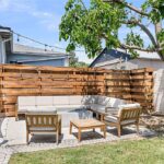 Backyard patio with curved sofa, wooden chairs, fence, and greenery.